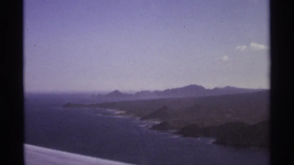 Aerial 8mm frame of the Cabo San Lucas coast, with blue ocean, rocky shore, mountains, and soft film color shift.
