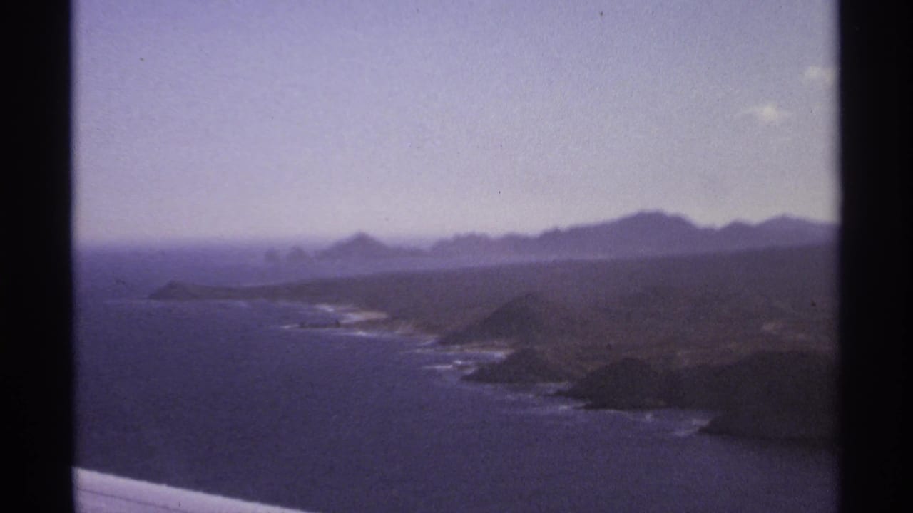 Hazy coastline with blue ocean, rocky headlands, distant hills, and dark film-frame edges.