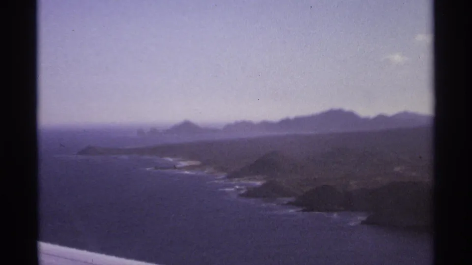 1974 8mm view of the Cabo San Lucas coastline with ocean, rugged headlands, pale sky, and distant hazy mountains.