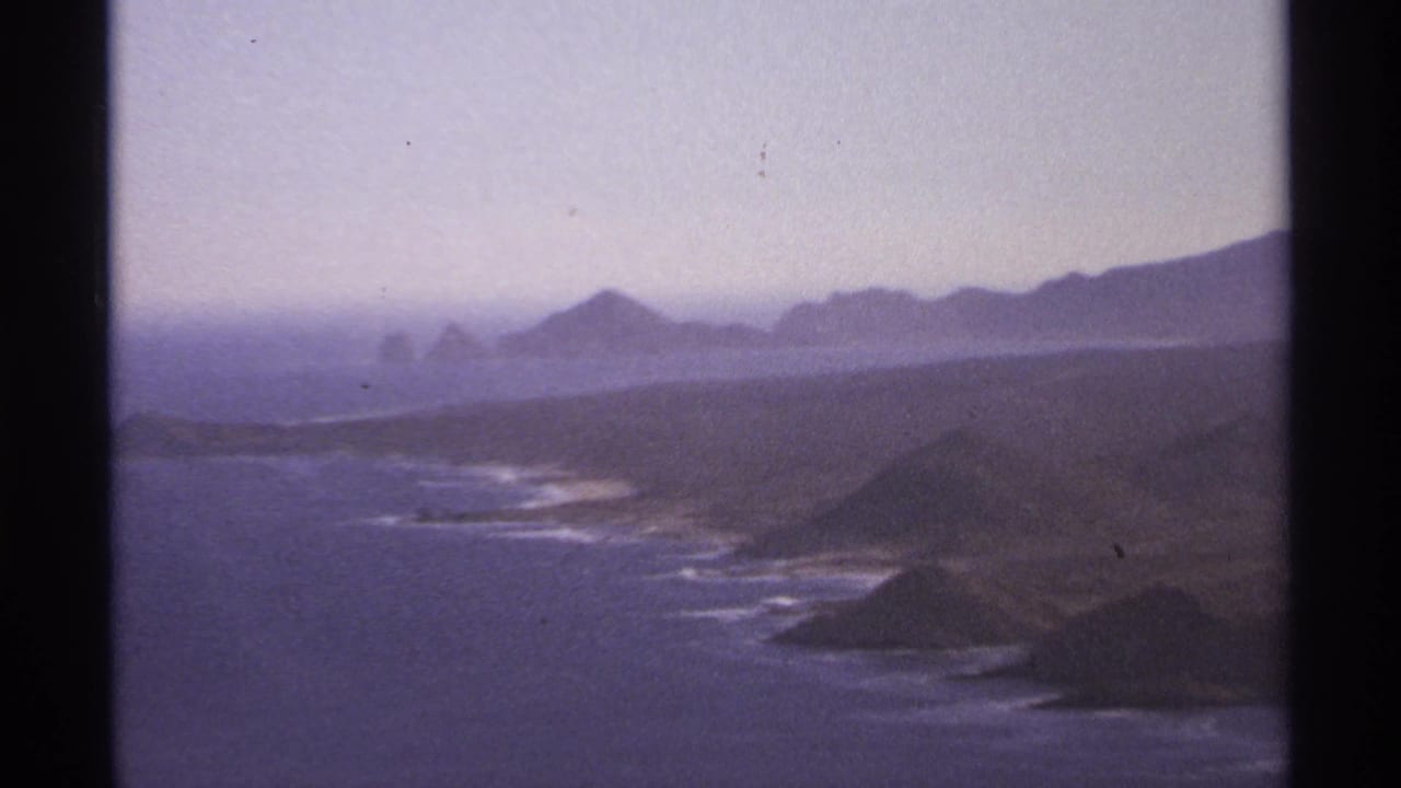 Ocean shoreline with rocky points, small islands, distant mountains, and pale hazy sky.