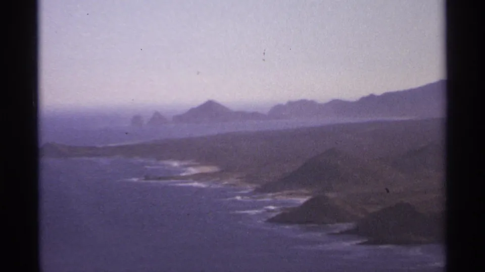 Cabo San Lucas coastal view on 8mm film, showing offshore rocks, rugged shoreline, ocean haze, and distant mountain forms.