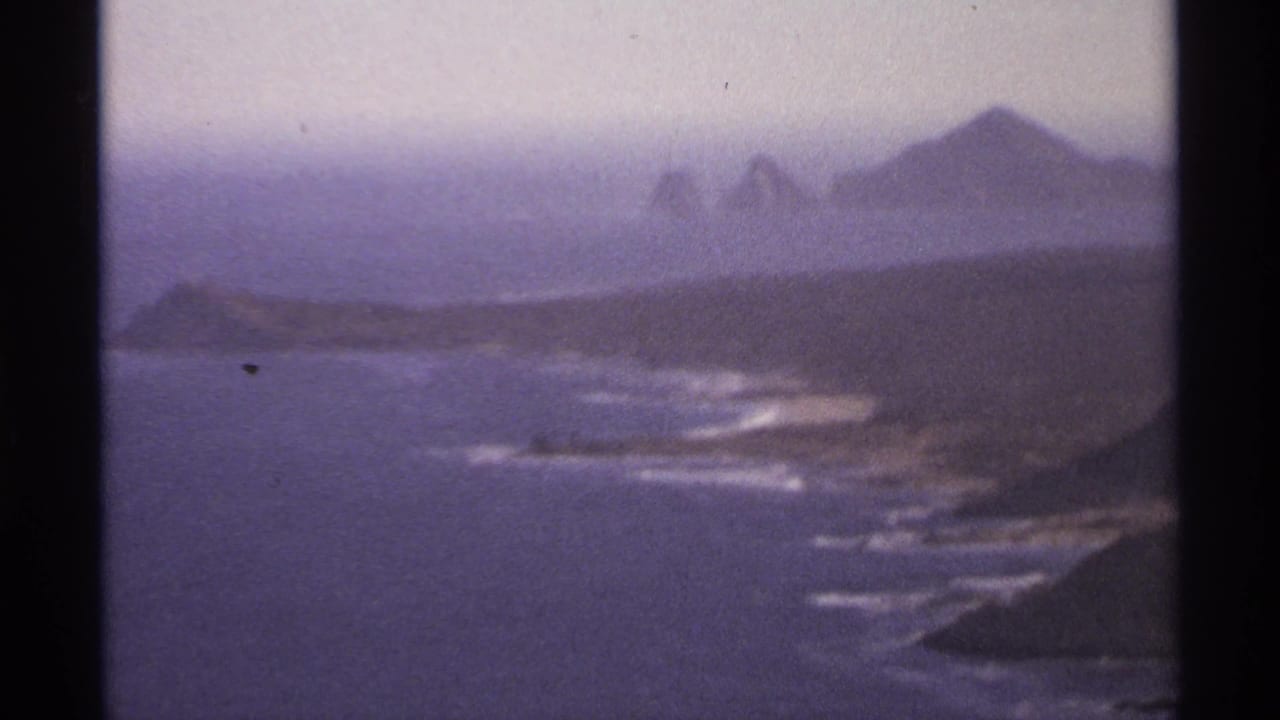 Hazy coastal shoreline with waves and distant rocky hills across the water.