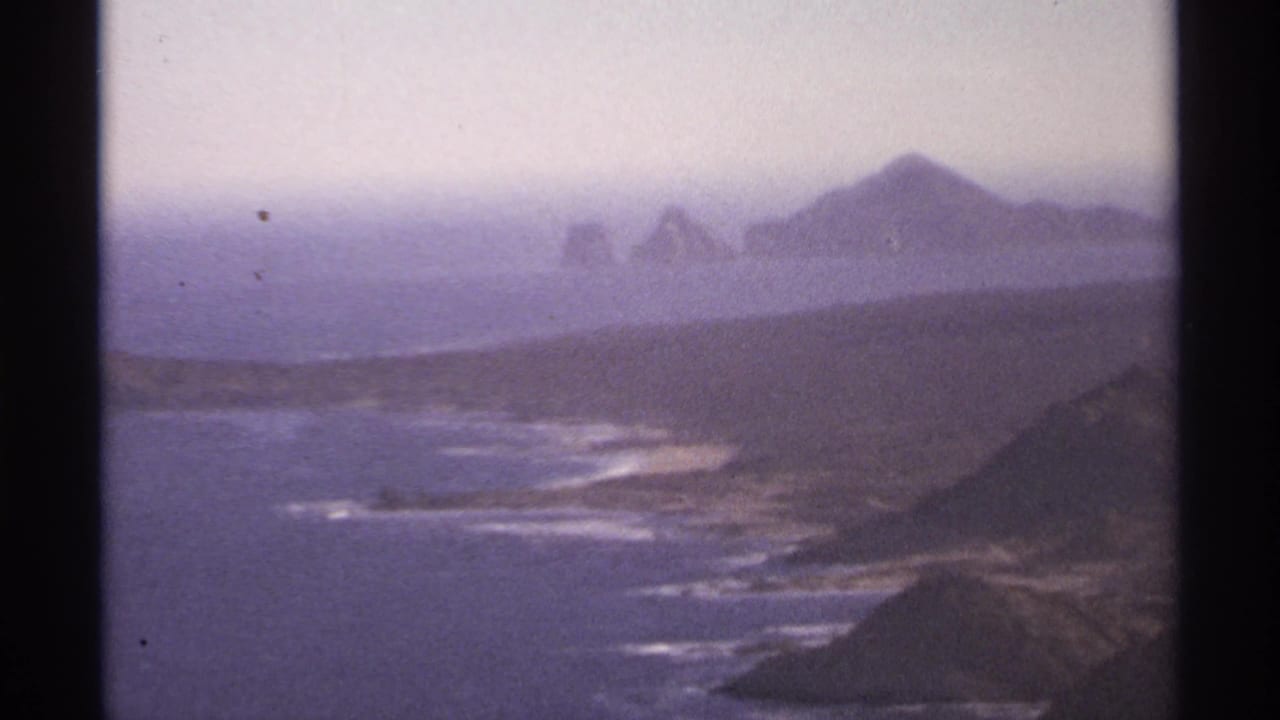 Coastline and surf seen from above, with distant rocky landforms on the horizon.
