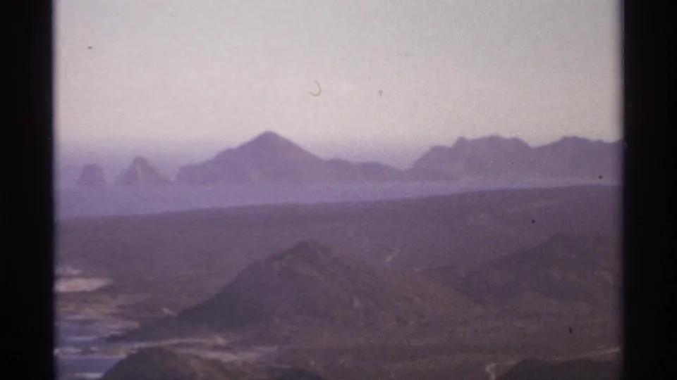 Soft 8mm view of the Cabo San Lucas coast in 1974, with dark hills, surf, and distant rocky island shapes.
