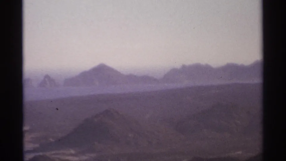 1974 Cabo San Lucas home-movie frame showing hazy mountain ridges and distant ocean through dark 8mm frame borders.