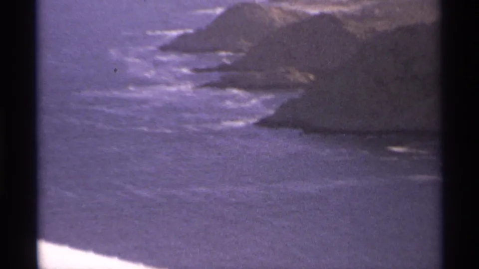 1974 8mm home-movie frame of Cabo San Lucas coastline, showing rocky headlands, surf, and purple-shifted film color.