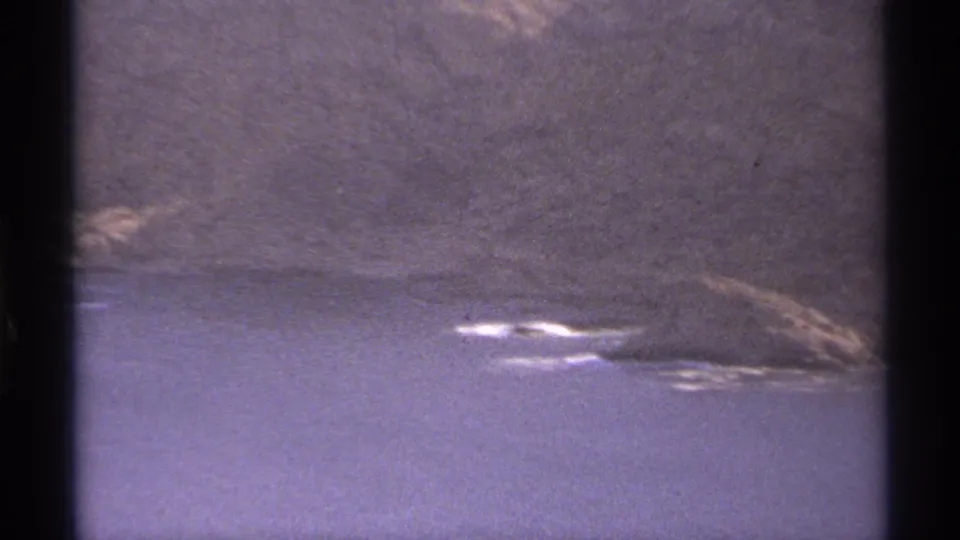 1974 Cabo San Lucas aerial view with blue water below a steep arid hillside, seen through the edges of an 8mm frame.