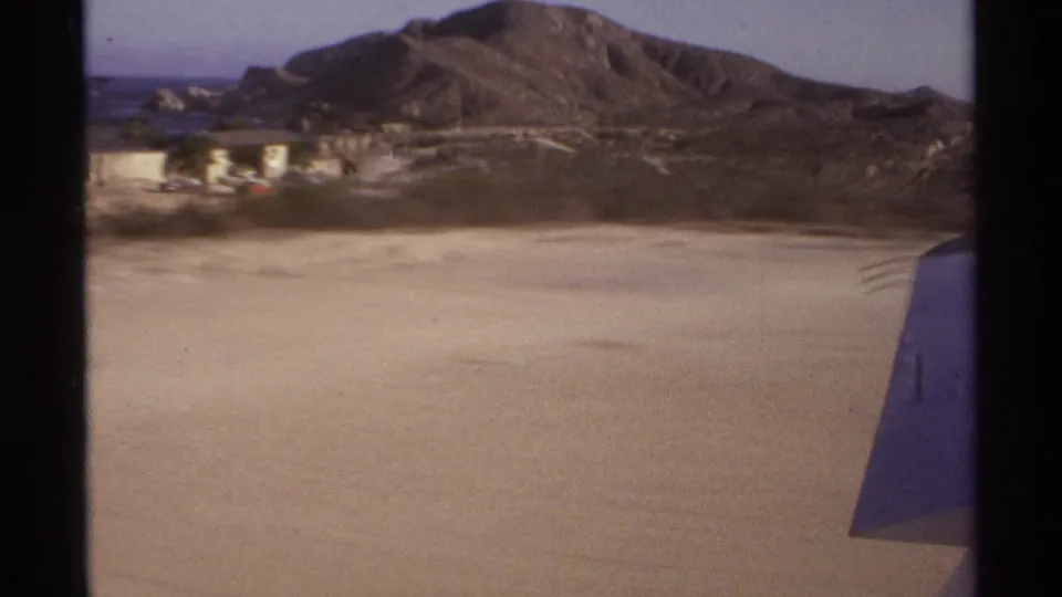 1974 aerial home-movie frame showing Cabo San Lucas hills, pale open ground, small structures, ocean at left, and a plane wing.