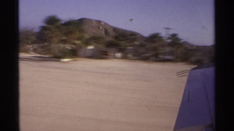 1974 Cabo San Lucas home-movie view from an aircraft window, showing the wing, runway, trees, and arid mountains.