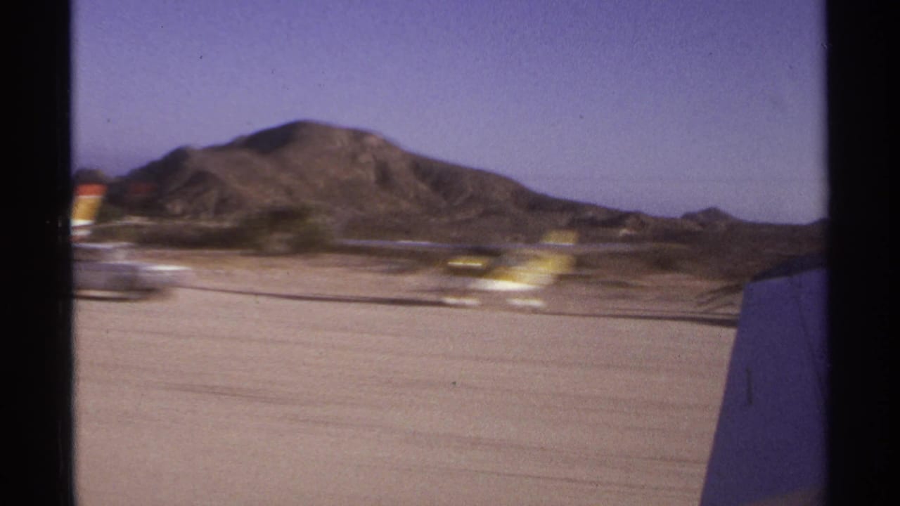 Blurred runway scene with aircraft wing, mountains, and small ground structures.