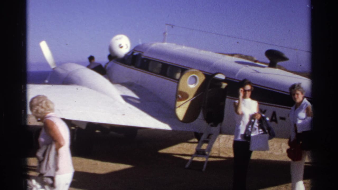 Two women stand near a small white propeller plane on a sunny airfield.