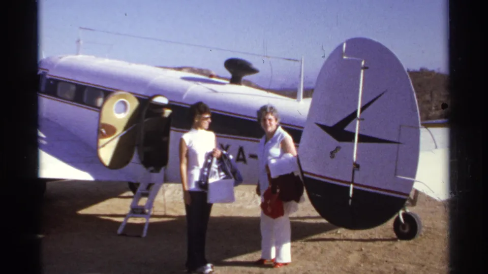 The 1974 home-movie frame shows two women posing beside a small white aircraft with a round tail and open passenger door.