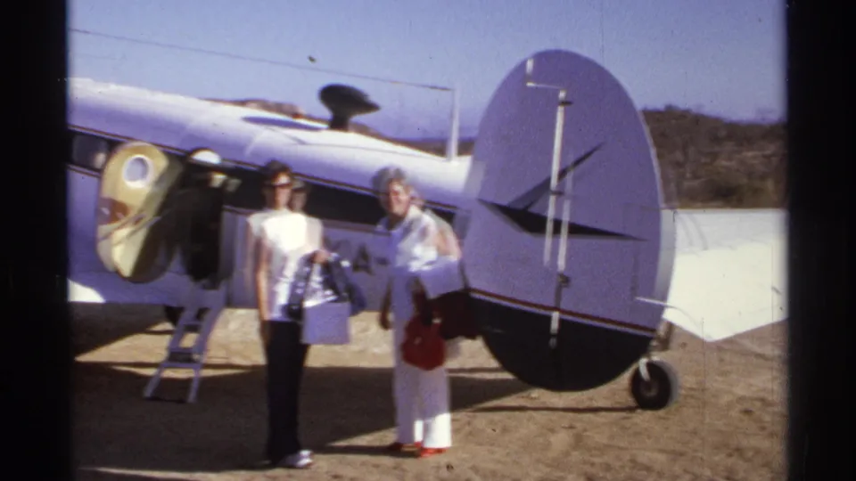 Two women stand by the tail and open door of a small aircraft in Cabo San Lucas, with 8mm softness and bright sun exposure.