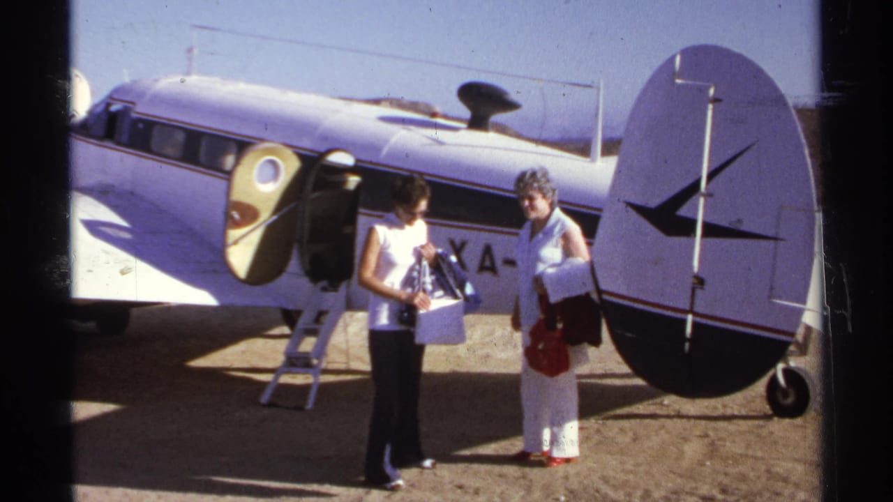 Two women stand beside a white propeller airplane with an open door on a sunny tarmac.