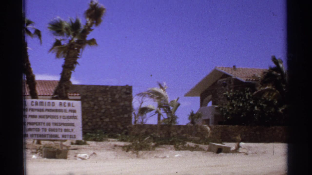 Hotel sign partly centered between palms, stone wall, resort building, sand, and sky.