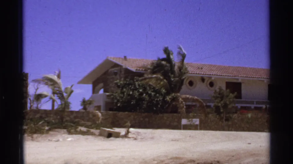 1974 Cabo San Lucas view of a resort building behind a stone wall with palms, sandy ground, and a small roadside sign.