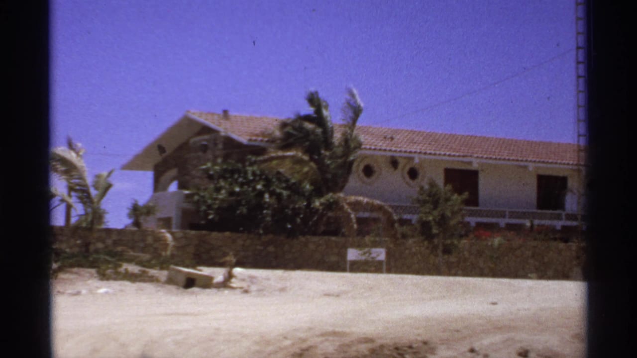 1974 Cabo San Lucas 8mm frame showing a sunlit beach house with a terracotta roof, palm trees, sandy ground, and dark frame edges.