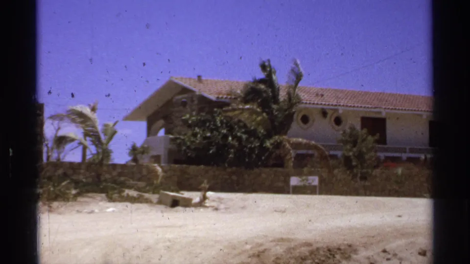 1974 Cabo San Lucas home-movie frame of a low beach house behind a wall, with palm trees moving in bright coastal light.