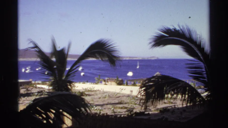 1974 Cabo San Lucas home-movie frame showing palm silhouettes and a window-like border over an ocean view with boats and distant shoreline.