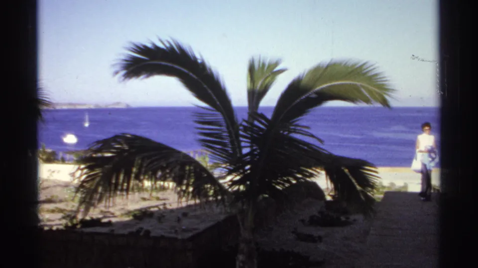 In 1974 Cabo San Lucas, an adult woman walks near a sandy beach path beneath palms with the ocean beyond.