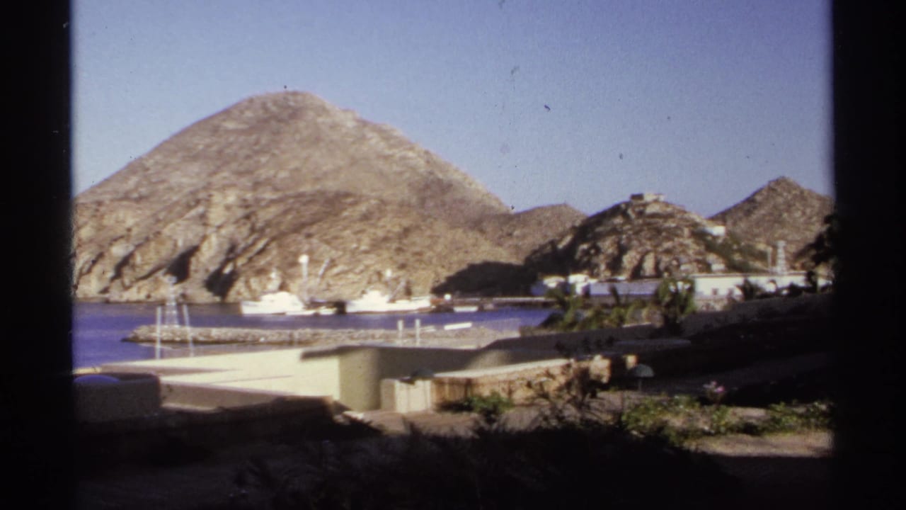 1974 Cabo San Lucas coastal frame with rocky hills above a harbor, small boats on blue water, low structures, and dark foreground framing.