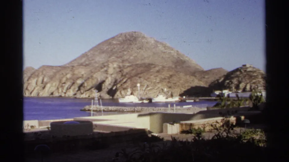 1974 Cabo San Lucas home-movie view across a harbor toward rocky hills, with boats, docks, low waterfront structures, and deep foreground shadow.