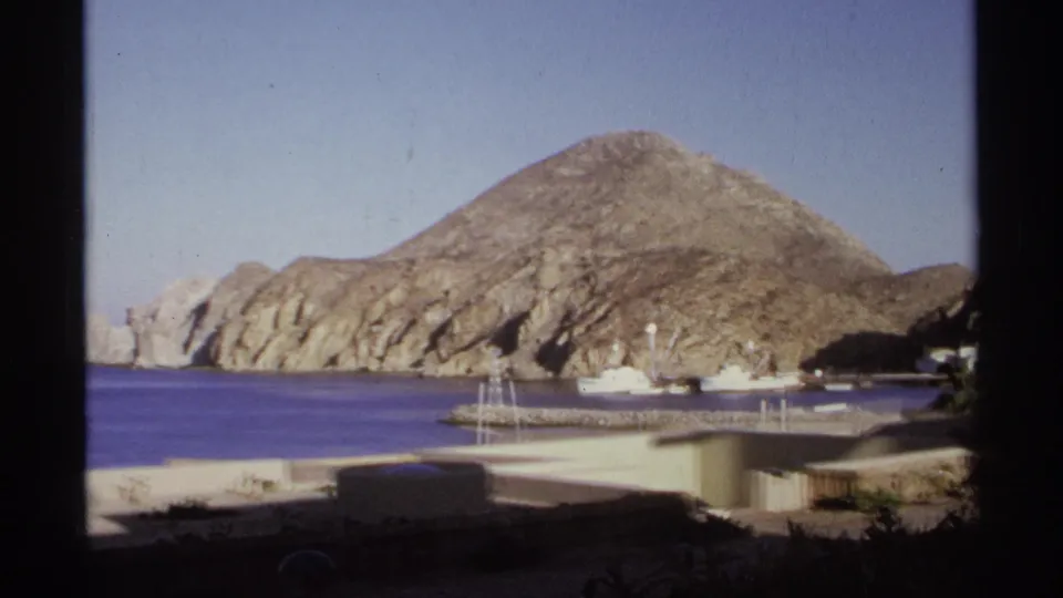 1974 Cabo San Lucas coastal frame showing rocky hills, harbor water, marina structures, a small boat, and dark foreground vegetation.