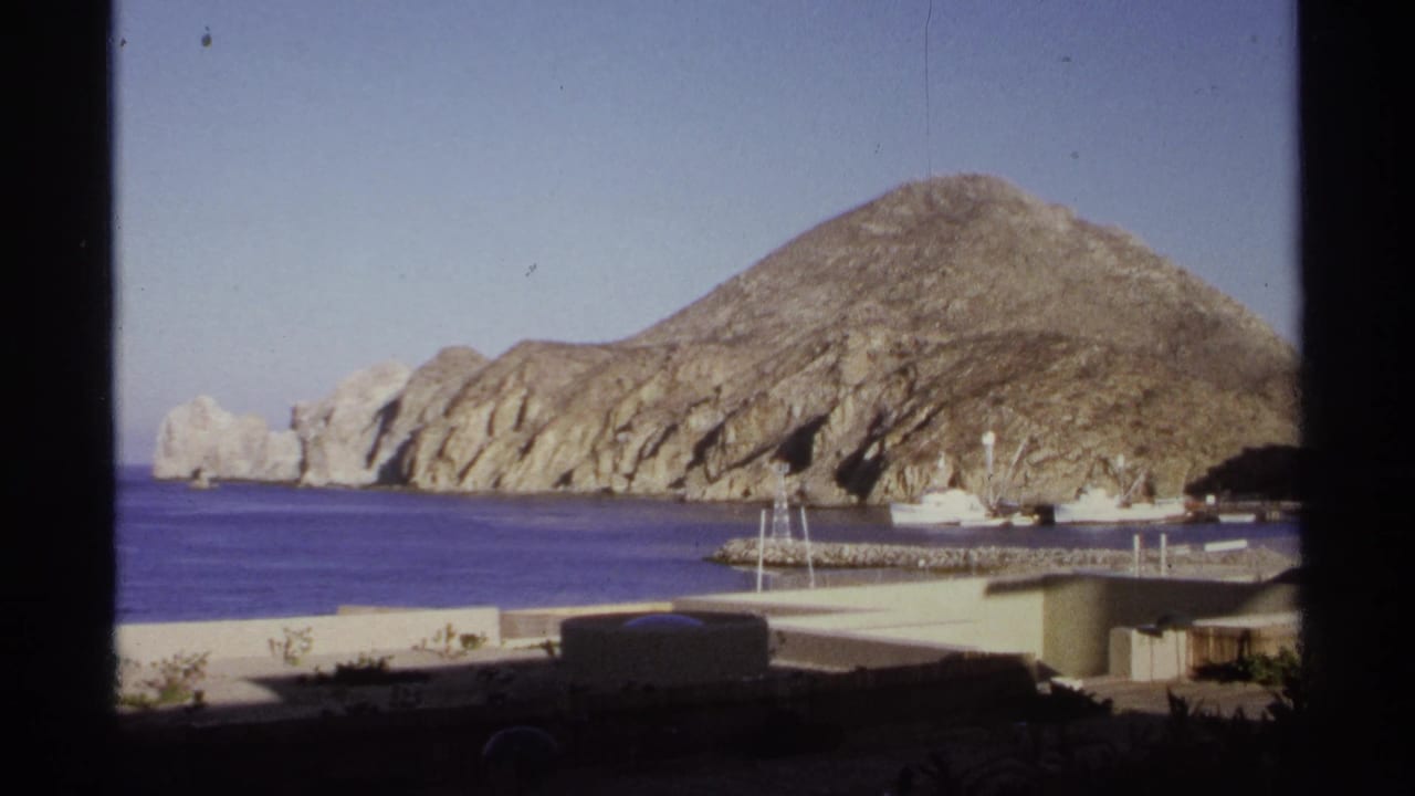 1974 Cabo San Lucas home-movie frame showing tan rocky coastline, blue water, harbor structures, and a clear sky.