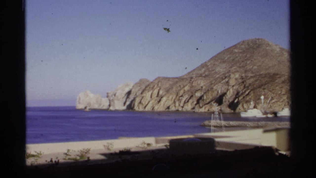 Blue bay, rocky tan headland, pale harbor edge, and small masts under a clear sky.