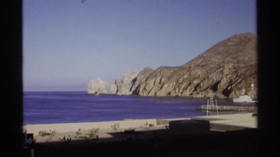 1974 Cabo San Lucas frame holding on rugged tan cliffs, calm blue ocean, small masts, and dark 8mm frame edges.