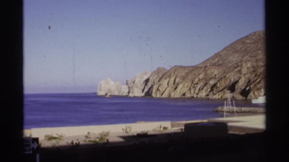 1974 Cabo San Lucas home-movie still showing tan rocky cliffs across blue water, with harbor masts and waterfront structures below.