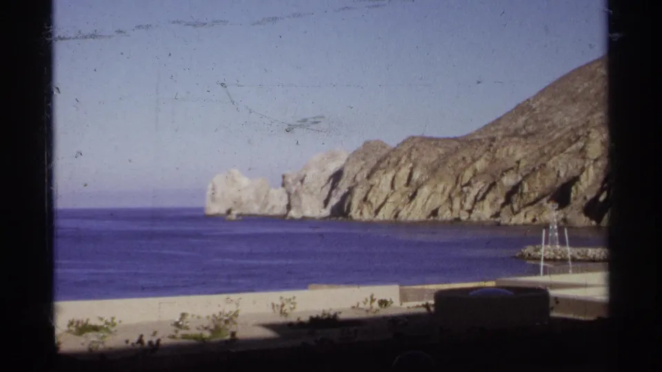 1974 Cabo San Lucas coastal view with rugged rocks, deep blue water, and a low wall near the shore.
