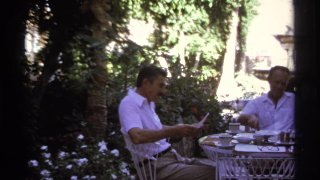 Two men seated at an outdoor table in a shaded garden.