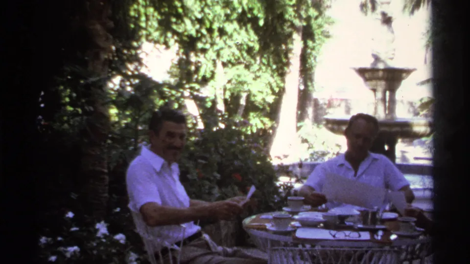 Two seated men at a white metal patio table, with papers, cups, foliage, and a fountain behind them.