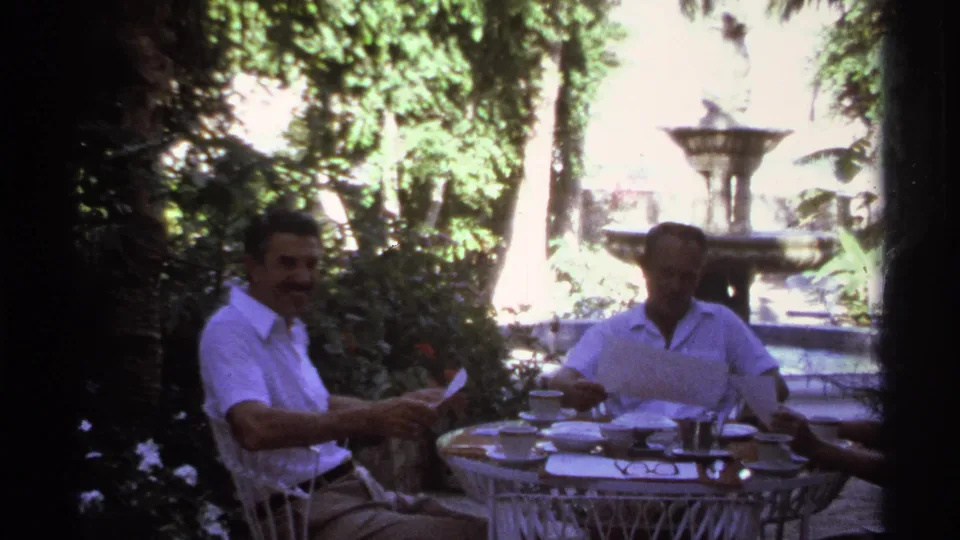 Two men sit at a patio table in a lush garden, with a decorative fountain and tableware visible behind and in front.