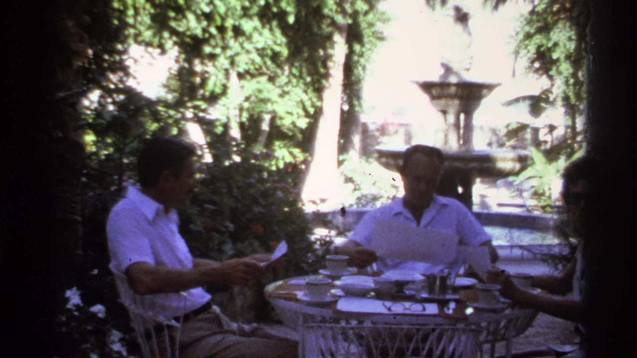 Three adults sit at a white wicker garden table with papers and dishes near a fountain.