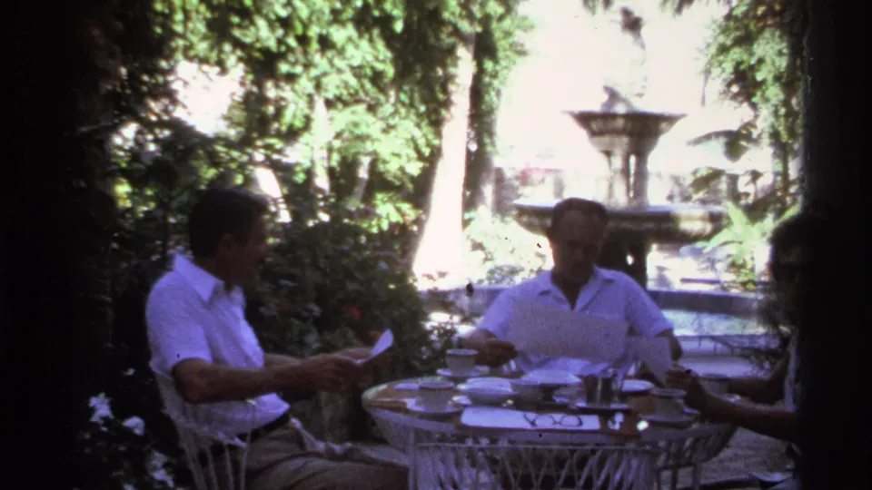 In 1974 Cabo San Lucas home-movie footage, three adults sit at a garden table with papers, dishes, foliage, and a stone fountain behind them.