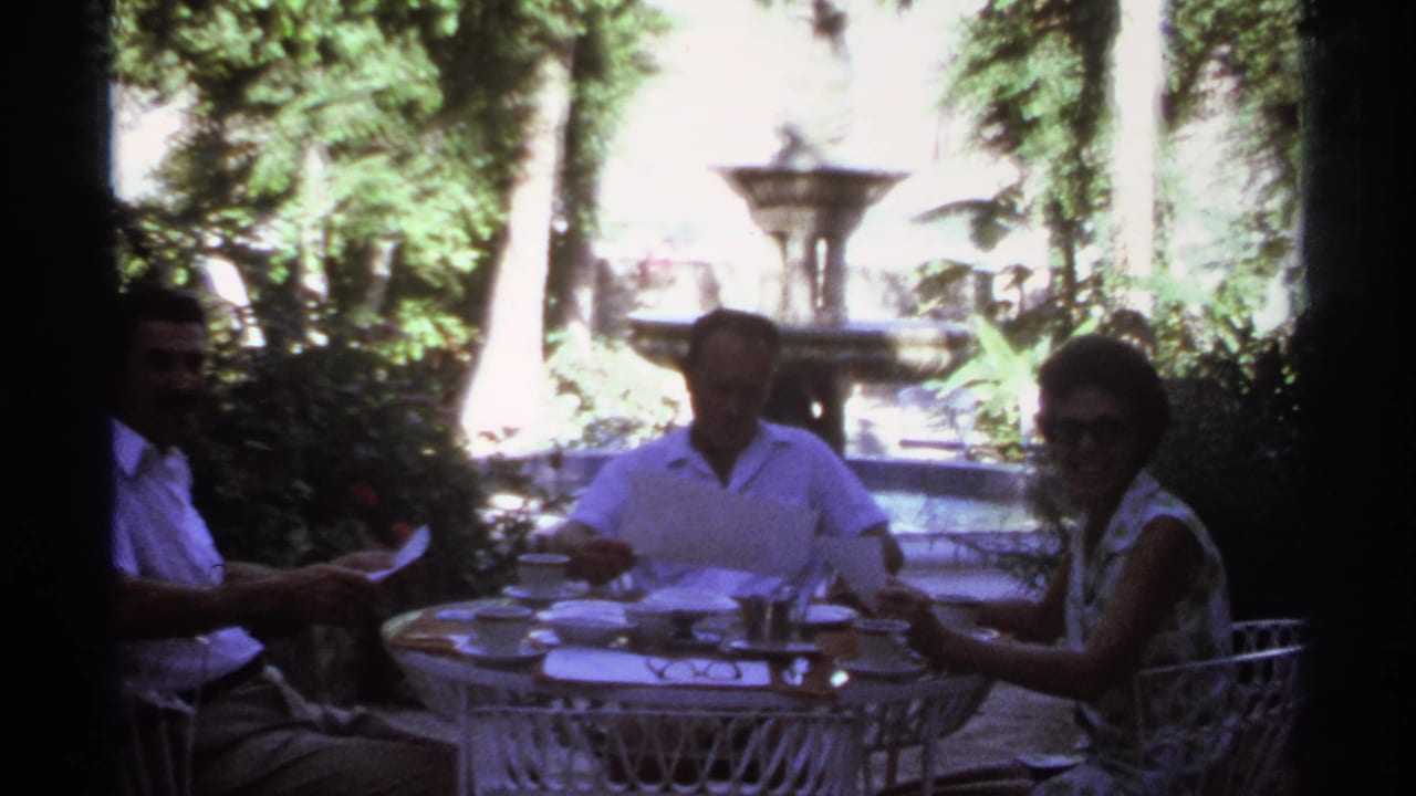 Adults sit around an outdoor table with papers while a woman in sunglasses faces the camera.