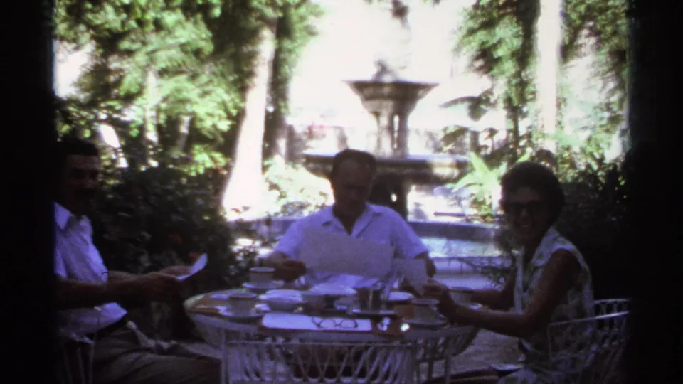 The family group remains seated at the garden table in Cabo San Lucas, with papers in hand, a woman in sunglasses, and a fountain in the background.