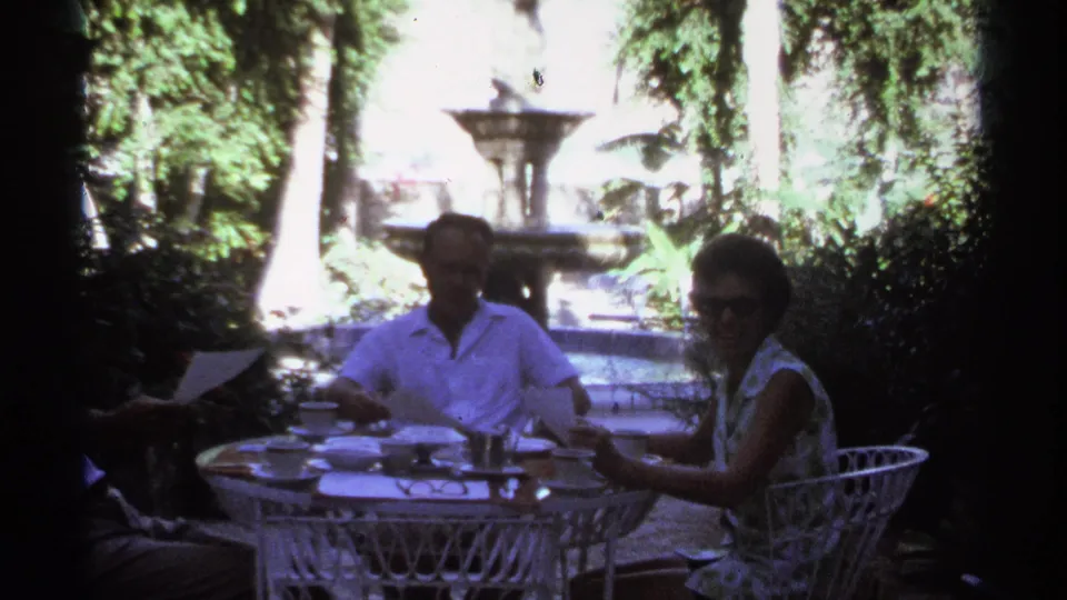 A 1974 Cabo San Lucas garden scene shows adults at a white wicker table, with one person partly cropped, papers on the table, and foliage around a fountain.