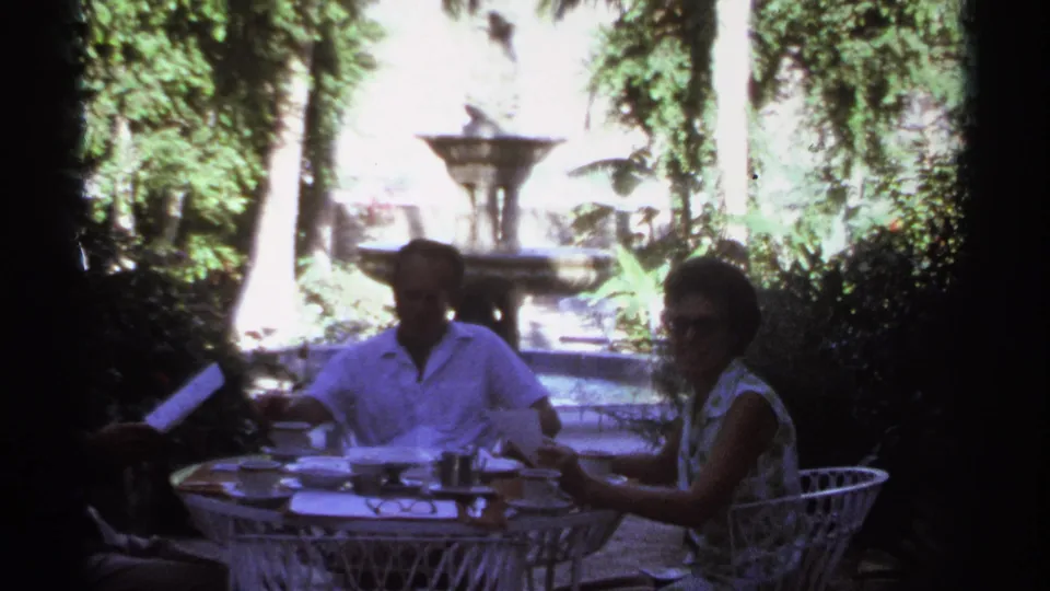 At the end of the Cabo San Lucas clip, a man and woman remain seated at the wicker table, with papers, dishes, foliage, and a fountain behind them.