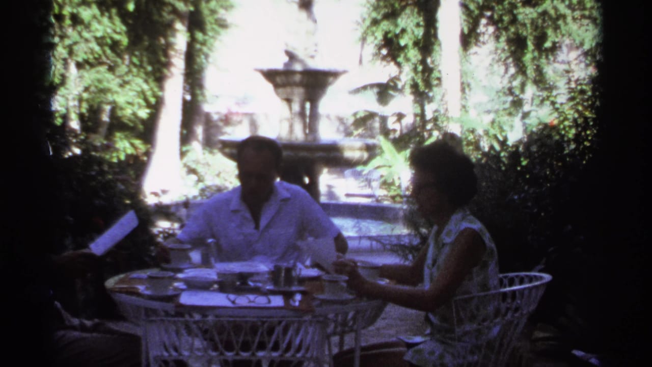 1974 Cabo San Lucas home-movie frame of adults at a white wrought-iron garden table with cups and a tiered fountain in the background.
