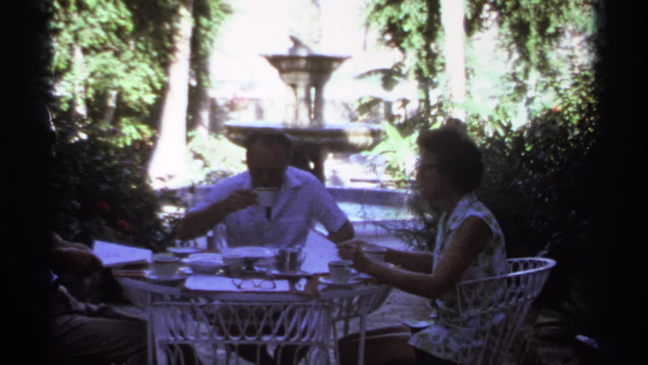 Man drinks from a cup while a woman sits across at a white patio table in a garden.