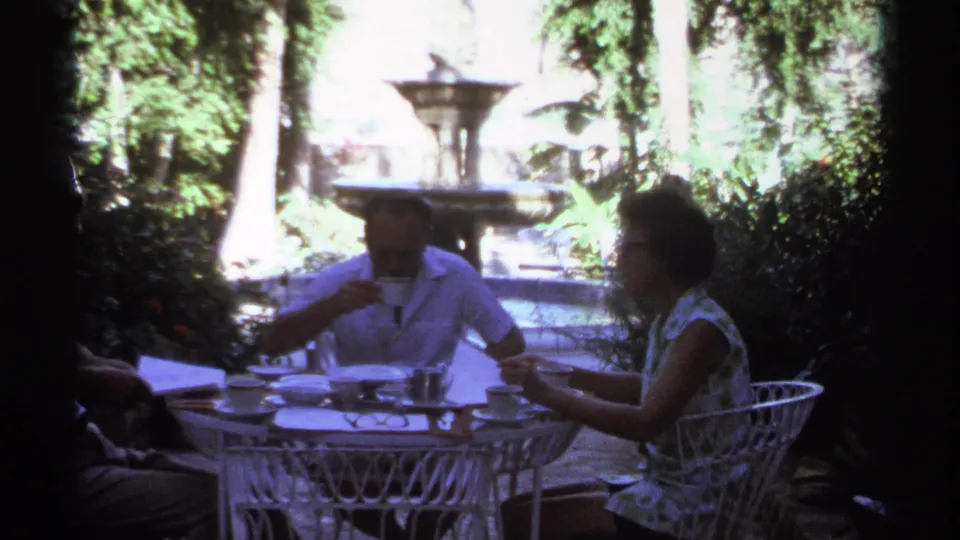 1974 Cabo San Lucas frame showing a man sipping from a cup beside a white patio table as a woman sits opposite in a lush garden.