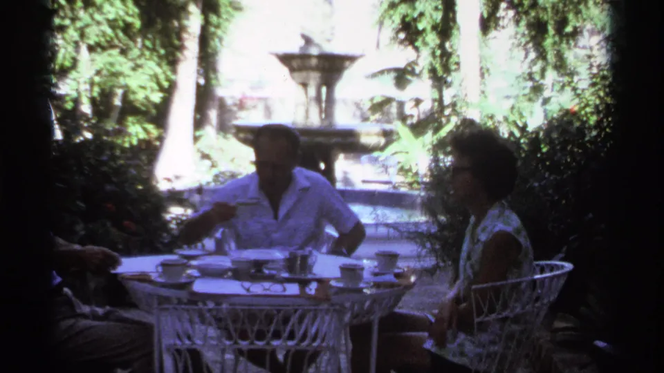 1974 Cabo San Lucas home-movie frame of adults seated at a round white garden table with cups, deep shade, and a fountain beyond.