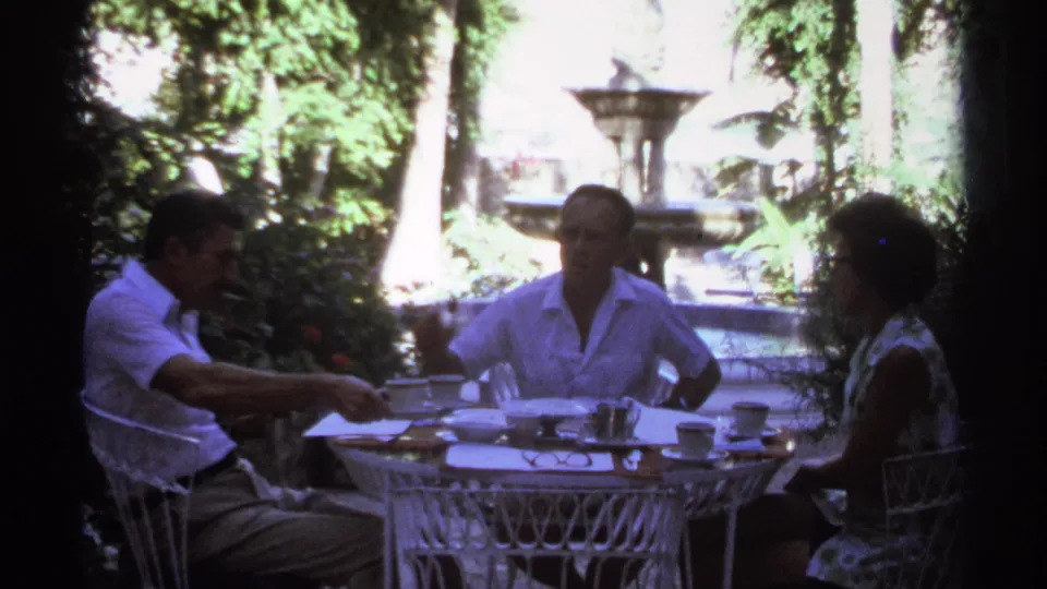 1974 Cabo San Lucas frame showing three adults gathered around a white wrought-iron table with cups, papers, greenery, and a fountain behind.