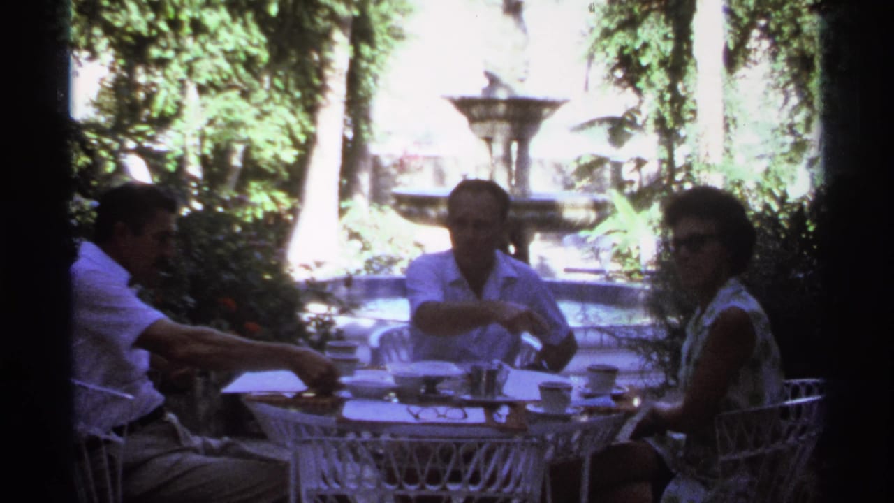 Three people seated at a white patio table in a garden with a fountain behind them.