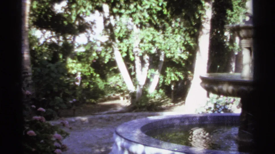 1974 home-movie view of a Cabo San Lucas garden, with a round fountain basin, path, palms, and shaded greenery.