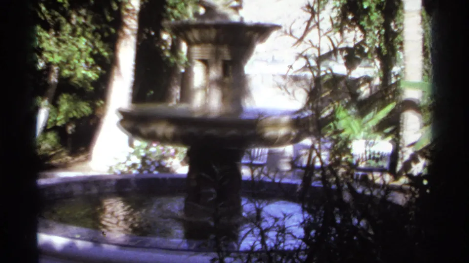 1974 Cabo San Lucas garden view with a stone fountain, circular basin, shaded trees, and sunlit tropical plants in soft 8mm color.