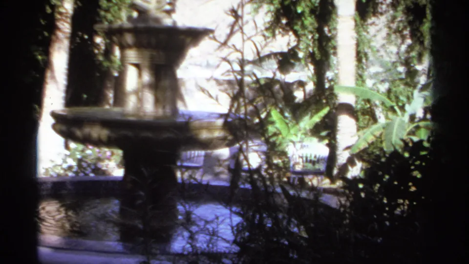 Soft 8mm frame from Cabo San Lucas in 1974 showing the fountain basin, tropical leaves, shrubs, and a garden bench or railing.